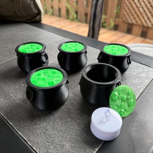 May include: A collection of miniature black cauldrons with bright green, bubbling contents, alongside a green, textured stone and a white LED tea light. The cauldrons have handles and are arranged on a dark surface, evoking a Halloween theme.