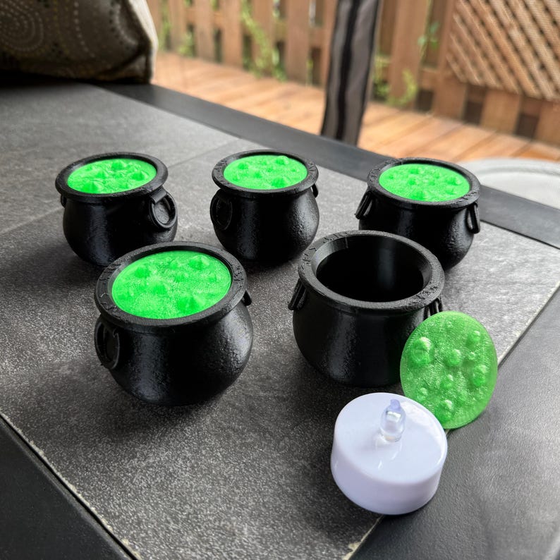 May include: A collection of miniature black cauldrons with bright green, bubbling contents, alongside a green, textured stone and a white LED tea light. The cauldrons have handles and are arranged on a dark surface, evoking a Halloween theme.