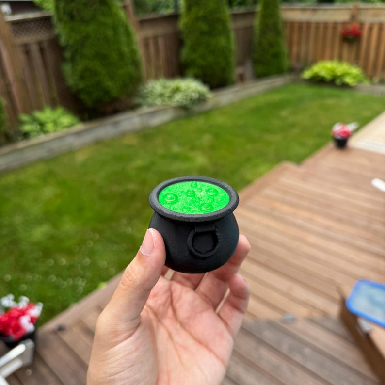 May include: A small, black cauldron with a green, bubbling liquid inside. The cauldron has a rounded shape with a thick rim and a small handle on the side. The green liquid appears to be glowing. The cauldron is held in a person's hand.