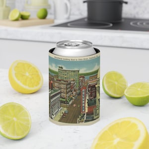 May include: A vintage-style can cooler featuring a postcard image of Broadway looking east to the Ohio River in Paducah, Kentucky. The image shows a street scene with buildings, cars, and a sign for the Columbia Theatre.