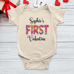 May include: A cream-colored baby onesie with the text "Sophie's FIRST Valentine" in black script. The word "FIRST" is filled with colorful heart shapes. The onesie is on a light wood-grain surface.