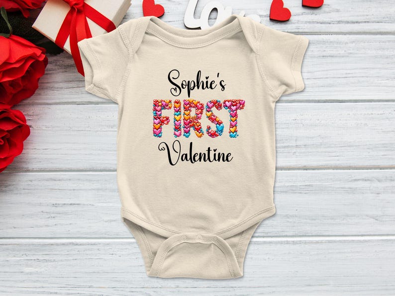 May include: A cream-colored baby onesie with the text "Sophie's FIRST Valentine" in black script. The word "FIRST" is filled with colorful heart shapes. The onesie is on a light wood-grain surface.