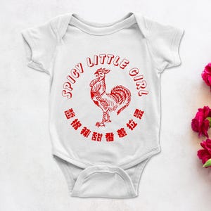 May include: White baby onesie with a red graphic of a rooster and the text "SPICY LITTLE GIRL" in a circular design. The onesie is on a white surface with pink flowers.