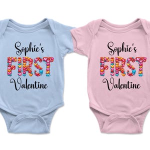 May include: Two baby onesies, one light blue and one pink. Each has the text "Sophie's FIRST Valentine" in black script. The word "FIRST" is made of colorful heart shapes, perfect for a Valentine's Day gift.