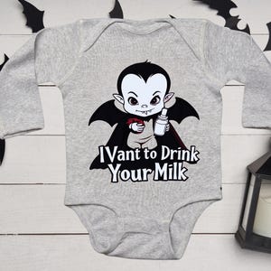 以下が含まれることがあります： 哺乳瓶を持った漫画の吸血鬼が描かれたグレーのベビーボディスーツ。吸血鬼は黒い翼と赤いマントをしています。「I vant to Drink Your Milk」の文字がプリントされています。装飾用のコウモリと黒いランタンが背景にあります。
