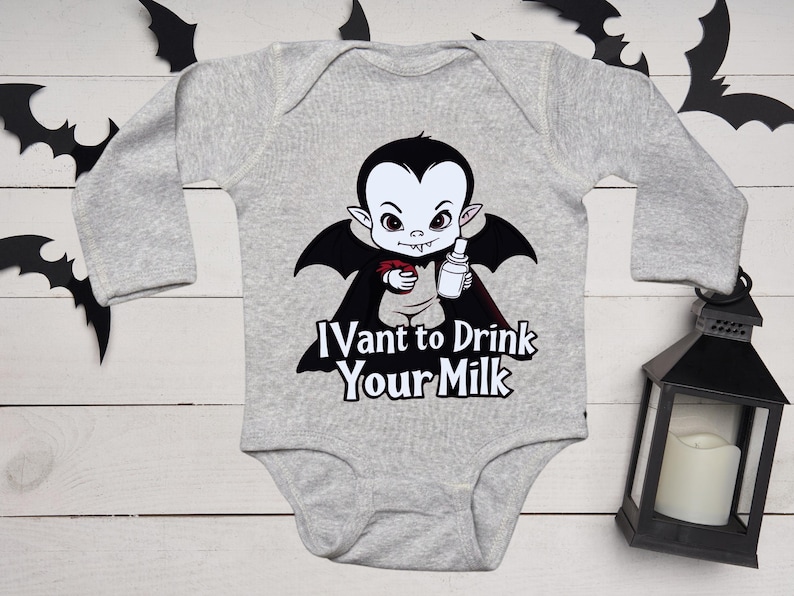 以下が含まれることがあります： 哺乳瓶を持った漫画の吸血鬼が描かれたグレーのベビーボディスーツ。吸血鬼は黒い翼と赤いマントをしています。「I vant to Drink Your Milk」の文字がプリントされています。装飾用のコウモリと黒いランタンが背景にあります。