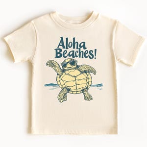 Pu&ograve; includere: T-shirt color crema con una tartaruga dei cartoni animati con occhiali da sole e la scritta turchese "Aloha Beaches!". La tartaruga &egrave; disegnata con le braccia tese e il disegno include un'onda blu.