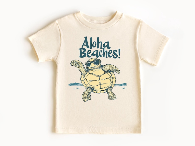 Pu&ograve; includere: T-shirt color crema con una tartaruga dei cartoni animati con occhiali da sole e la scritta turchese "Aloha Beaches!". La tartaruga &egrave; disegnata con le braccia tese e il disegno include un'onda blu.