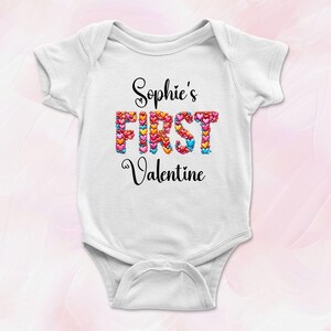 May include: White baby onesie with the text "Sophie's FIRST Valentine" in black script. The word "FIRST" is filled with colorful hearts. A cute outfit for a baby's first Valentine's Day.