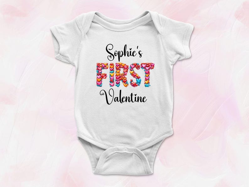May include: White baby onesie with the text "Sophie's FIRST Valentine" in black script. The word "FIRST" is filled with colorful hearts. A cute outfit for a baby's first Valentine's Day.