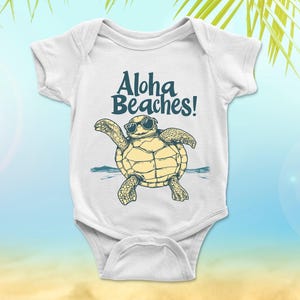 Pu&ograve; includere: Body da bambino bianco con la scritta "Aloha Beaches!" sopra una tartaruga dei cartoni animati con gli occhiali da sole. La tartaruga &egrave; gialla e verde, con acqua blu sullo sfondo. Il body ha maniche corte e chiusura a scatto.