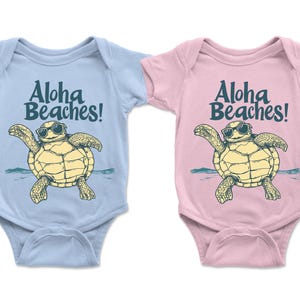 Pu&ograve; includere: Due body per neonati, uno azzurro chiaro e uno rosa, ciascuno con una tartaruga dei cartoni animati con occhiali da sole. Il testo "Aloha Beaches!" &egrave; stampato sopra la tartaruga. La tartaruga &egrave; gialla e beige.