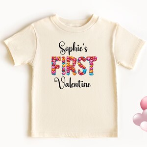 May include: Cream-colored t-shirt with the text "Sophie's FIRST Valentine" in black script. The word "FIRST" is made of colorful heart shapes. A cluster of pink heart-shaped balloons is in the lower right corner.