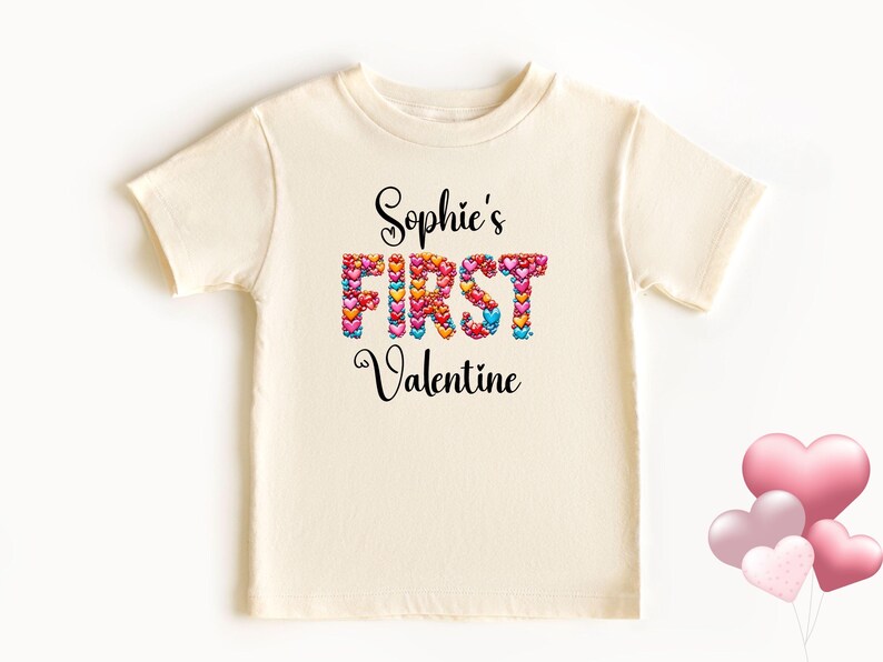 May include: Cream-colored t-shirt with the text "Sophie's FIRST Valentine" in black script. The word "FIRST" is made of colorful heart shapes. A cluster of pink heart-shaped balloons is in the lower right corner.