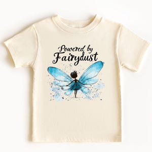 Puede incluir: Camiseta color crema con la frase "Powered by Fairydust" sobre una ilustraci&oacute;n de acuarela de un hada con alas azules. El hada tiene una silueta oscura y est&aacute; rodeada de salpicaduras azules.