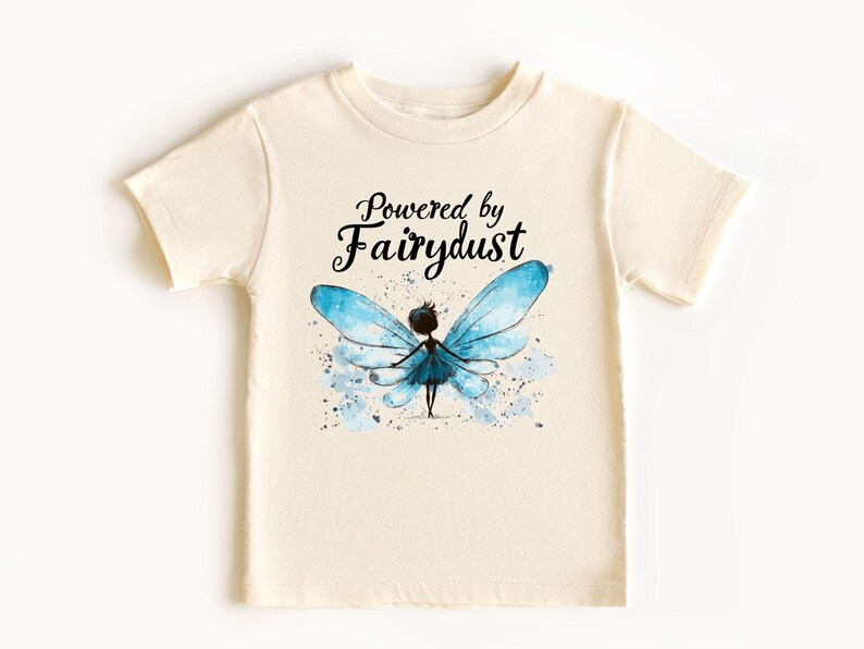 Puede incluir: Camiseta color crema con la frase "Powered by Fairydust" sobre una ilustraci&oacute;n de acuarela de un hada con alas azules. El hada tiene una silueta oscura y est&aacute; rodeada de salpicaduras azules.