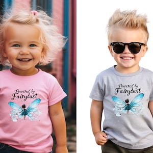 Puede incluir: Dos camisetas infantiles, una rosa y otra gris, con el texto "Powered by Fairydust" y un dise&ntilde;o de hada. El hada tiene alas azules y un vestido azul.