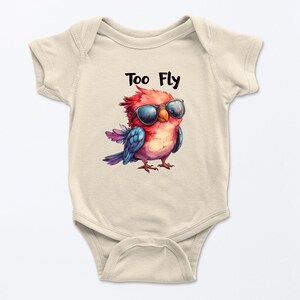 May include: A light beige baby onesie with a colorful illustration of a bird wearing sunglasses. The bird is red, blue, and purple, with the text "Too Fly" above it in black. The onesie is made of soft material.