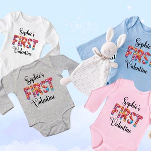 May include: Four baby onesies in white, gray, blue, and pink, each with the text "Sophie's FIRST Valentine." The word "FIRST" is made of colorful hearts. A stuffed bunny and teddy bear are also in the image.