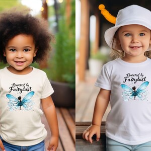 Puede incluir: Dos ni&ntilde;os con camisetas blancas con el texto "Powered by Fairydust" y un dise&ntilde;o de hada azul. Las camisetas son de manga corta. Un ni&ntilde;o tiene el pelo rizado oscuro, y el otro tiene el pelo rubio y lleva un sombrero blanco.