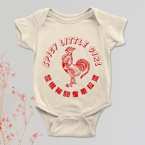 May include: Cream-colored baby onesie with a red rooster graphic and the words "SPICY LITTLE GIRL" in a circular design. The onesie also features Chinese characters. A cute and stylish outfit for a baby.