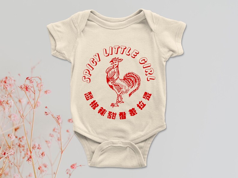 May include: Cream-colored baby onesie with a red rooster graphic and the words "SPICY LITTLE GIRL" in a circular design. The onesie also features Chinese characters. A cute and stylish outfit for a baby.