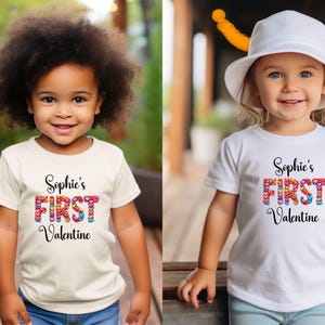 May include: Two young children wearing white and cream-colored t-shirts. The shirts have the text "Sophie's FIRST Valentine" in a colorful heart design. The children are smiling and wearing blue jeans.