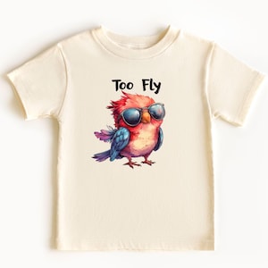 May include: Cream-colored t-shirt featuring a colorful cartoon bird wearing sunglasses. The bird is red, blue, and pink, with the text "Too Fly" above it in black. The shirt is made of a soft material.