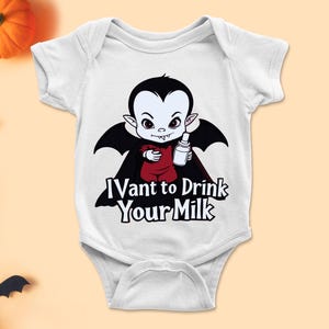 以下が含まれることがあります： 白いベビーボディスーツ。黒いマントを着て哺乳瓶を持った漫画の吸血鬼が描かれています。「I Vant to Drink Your Milk」の文字がプリントされています。ハロウィーンに最適です。