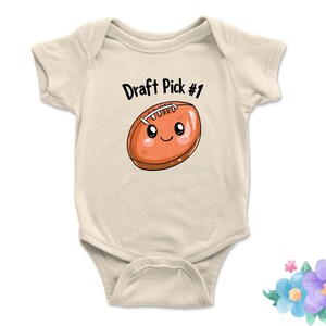Puede incluir: Body de bebé color crema con el texto "Draft Pick #1" y un balón de fútbol de dibujos animados con una cara sonriente. El balón es marrón con cordones blancos y un contorno marrón claro.