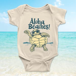Pu&ograve; includere: Body da bambino color crema con una tartaruga dei cartoni animati con occhiali da sole. La tartaruga &egrave; gialla e verde, con le parole "Aloha Beaches!" in blu navy sopra. Lo sfondo mostra una spiaggia e l'oceano.