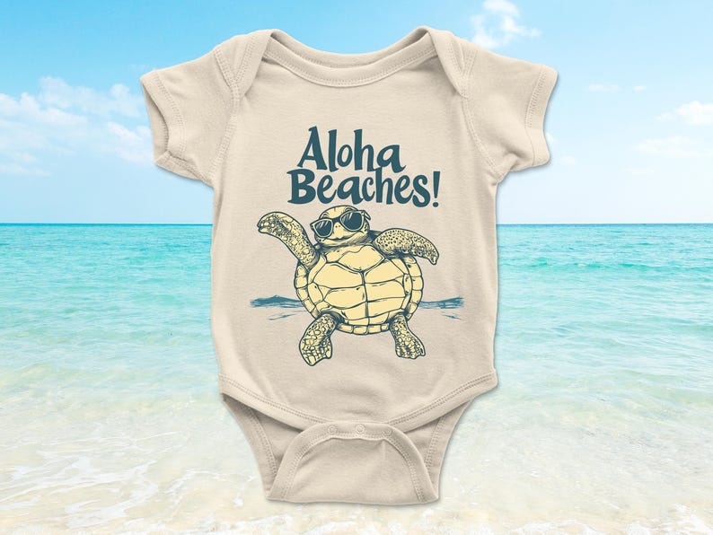 Pu&ograve; includere: Body da bambino color crema con una tartaruga dei cartoni animati con occhiali da sole. La tartaruga &egrave; gialla e verde, con le parole "Aloha Beaches!" in blu navy sopra. Lo sfondo mostra una spiaggia e l'oceano.