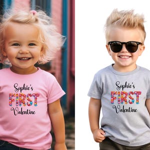 May include: Two children wearing t-shirts with the text "Sophie's FIRST Valentine". The text is in black script with the word "FIRST" in colorful heart shapes. One shirt is pink, the other is gray.