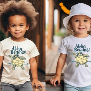 Pu&ograve; includere: Due bambini con magliette bianche e color crema. Le magliette presentano una tartaruga dei cartoni animati con occhiali da sole con la scritta "Aloha Beaches!" in blu. I bambini sorridono e guardano la fotocamera.