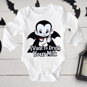 以下が含まれることがあります： 白いベビーボディスーツ。漫画の吸血鬼のデザイン。黒いマントを着た吸血鬼が、ボトルと小さな赤い物体を持っています。ボディスーツのテキストは「I vant to drink your milk」です。黒いコウモリの切り抜きと黒いランタンが背景にあります。
