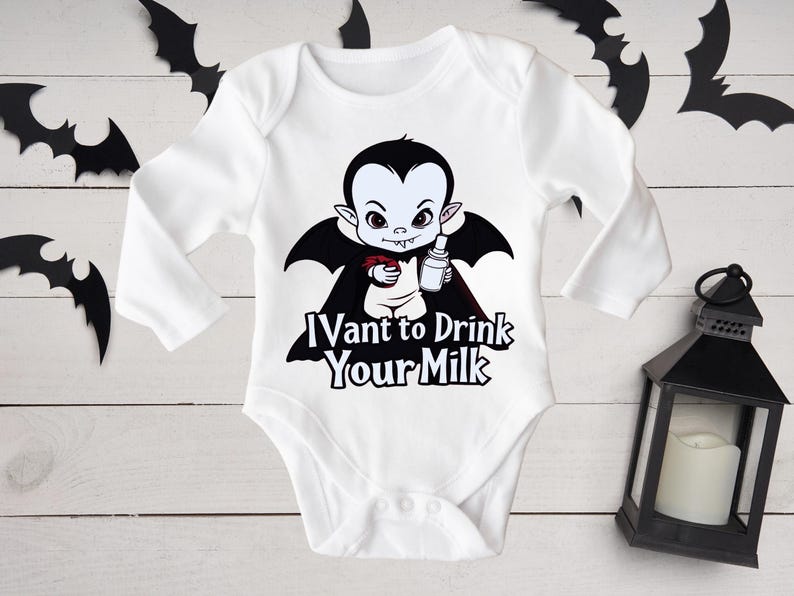 以下が含まれることがあります： 白いベビーボディスーツ。漫画の吸血鬼のデザイン。黒いマントを着た吸血鬼が、ボトルと小さな赤い物体を持っています。ボディスーツのテキストは「I vant to drink your milk」です。黒いコウモリの切り抜きと黒いランタンが背景にあります。