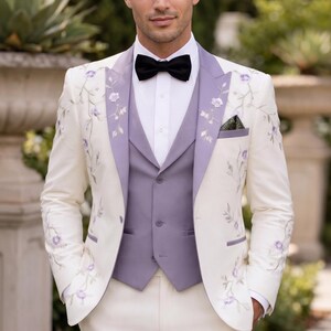 May include: Ivory suit with floral embroidery, featuring a lavender vest and lapel trim. The ensemble includes a black bow tie and a pocket square. The suit is a formal wear option.