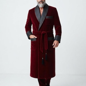 May include: A deep burgundy velvet robe featuring a black quilted shawl collar, matching black quilted cuffs, and a black quilted chest pocket. The robe includes a matching velvet tie belt with black tassels and coordinating burgundy velvet trousers.