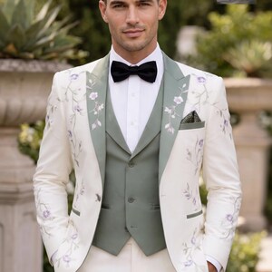 May include: Ivory suit with floral embroidery, featuring a sage green vest and lapels, paired with a black bow tie. The suit includes a matching pocket square. The image displays the "RUBEE FASHION" logo.