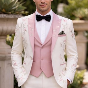 May include: A three-piece cream suit with floral embroidery, featuring a pink vest and lapel trim, paired with a black bow tie. The suit is worn with a white dress shirt and matching cream trousers. The ensemble is suitable for formal events.