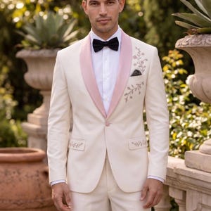May include: A cream-colored suit jacket featuring a pink shawl lapel, a black bow tie, and a white button-down shirt. The jacket includes delicate floral embroidery on the left chest and pocket flaps, with the Rubee Fashion logo visible.