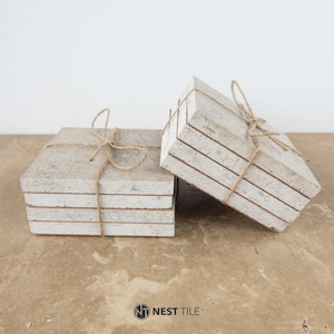 May include: Two stacks of square stone coasters tied with twine. The coasters are a light beige color with visible veining, suggesting a natural stone material. The twine adds a rustic touch to the presentation. The coasters are likely around 4 inches square.