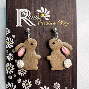 May include: A pair of brown bunny rabbit earrings with pink ears and white fluffy tails. The earrings are on a brown wooden display with a white daisy pattern and the text "Rae's Creative Clay".