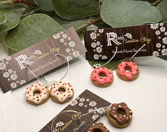 May include: A pair of donut-shaped earrings with pink and white frosting and sprinkles, and another pair with chocolate and white frosting and sprinkles. The earrings are handmade polymer clay jewelry and are displayed on a wooden background with a white daisy pattern.