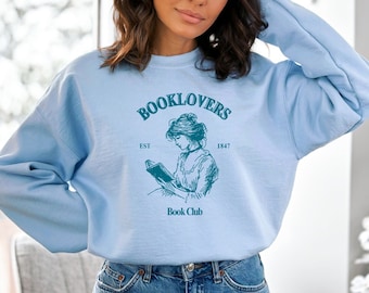 May include: A light blue sweatshirt with a graphic of a woman reading a book. The graphic includes the text "Booklovers Est 1847 Book Club".