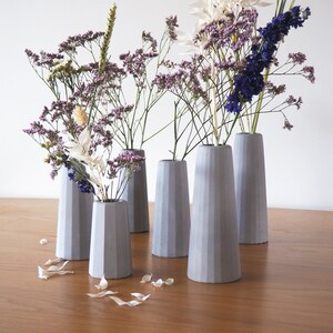 May include: Six gray concrete vases of varying heights with a variety of dried flowers in them. The vases are all textured and have a modern design.