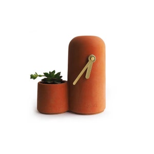 Può includere: Un orologio in terracotta color terracotta con una lancetta dorata e un piccolo vaso in terracotta con una pianta grassa. L'orologio ha la scritta "done's" stampata sul lato.