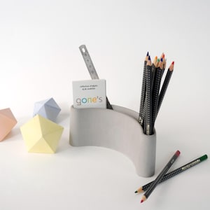 Peut inclure: Un organiseur de bureau en béton gris avec un design incurvé, contenant des crayons, une règle et une carte de visite. Des sculptures géométriques en papier aux couleurs pastel sont dispersées autour. La carte de visite indique "gone's".
