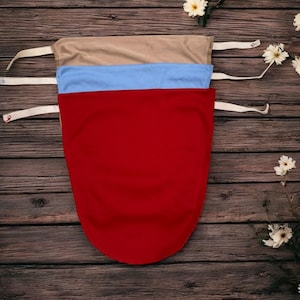 May include: Three fabric face coverings in red, light blue, and tan, each with white straps. The coverings are stacked on a dark wood surface, with white flowers scattered around.