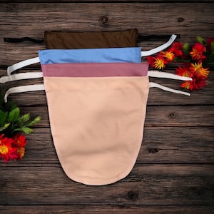 Könnte beinhalten: Vier Stoff-Gesichtsbedeckungen in Braun, Blau, Mauve und Pfirsich. Jede hat einen gebogenen Boden und elastische Träger. Die Bedeckungen sind auf einer dunklen Holzoberfläche gestapelt, mit roten und gelben Blumen im Hintergrund.